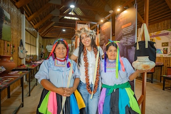 Tres mujeres posan en un salón con paredes y techo de madera. Una mujer lleva tocado de plumas; las otras dos, atuendos indígenas y una sostiene una vasija