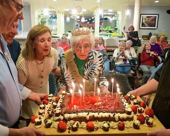 Una mujer mayor con tiara y banda apaga las velas de un pastel grande decorado con fresas frente a un grupo de personas