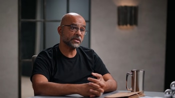 Mo Gawdat, un hombre calvo con barba y gafas, viste una camiseta negra mientras habla. Está sentado frente a una mesa con una taza y papeles
