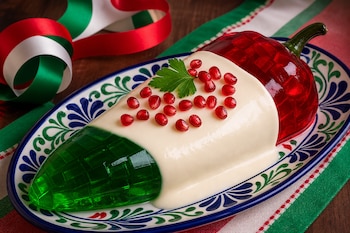 Una gelatina con los colores verde, blanco y rojo sobre un plato decorado, acompañada por granada, perejil y listones tricolores.