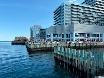 Edificios modernos con balcones, muelle de madera con pilotes, personas sentadas, mar azul con olas, cielo despejado, mesas, sillas, sombrillas