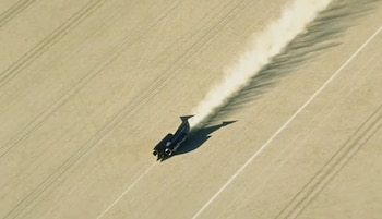 Vehículo negro aerodinámico con dos motores traseros, estela de polvo blanco, terreno claro y plano con líneas diagonales