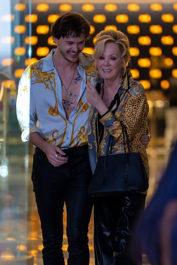 Christopher Briney, de camisa abierta azul y dorada, y Jean Smart, de blusa estampada, sonríen. Ambos visten pantalones negros, ella lleva bolso negro