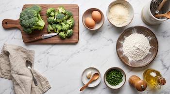 Brócoli, huevos, harina, queso rallado, ajo, perejil, aceite y utensilios organizados sobre una superficie de mármol blanco.