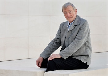 Hombre senior de cabello gris y chaqueta gris sentado contra una pared de azulejos blancos, mirando ligeramente a la cámara con una sonrisa tenue