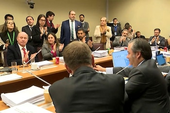 Marinos Kartikkis y Jorge Quiroz, vestidos de traje, se dan la mano y sonríen a la cámara en una sala grande con decoraciones doradas y medios de comunicación