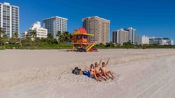 Tres personas en traje de baño sentadas en una playa de arena clara, con una torre de salvavidas roja y amarilla, palmeras y edificios altos bajo un cielo azul