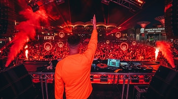 Un DJ de espaldas con suéter naranja maneja una consola frente a la multitud bajo luces rojas y un letrero del Parque Ecológico Aragón.