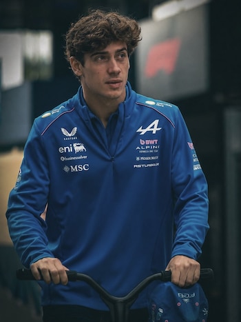 Un joven de cabello rizado con sudadera azul de Alpine, sosteniendo manillares negros con ambas manos. Un gorro azul está colgado