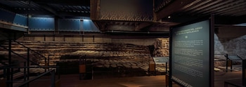 Estructuras de piedra antiguas, escalones de mampostería, barandas de metal, techo oscuro con vigas y dos paneles informativos con texto en una exposición