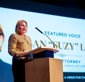Mujer rubia con chaqueta dorada habla en un podio de madera con micrófono. Pantalla de fondo muestra texto "Featured Voice", "Susan "Suzy" Lo", "Attorney", "Circuit of Florida"