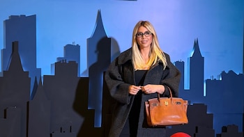 Wanda Nara, mujer rubia con gafas y abrigo oscuro a cuadros, posa frente a un fondo azul con siluetas de ciudad y el cartel gigante de 'Annie'. Lleva un bolso marrón