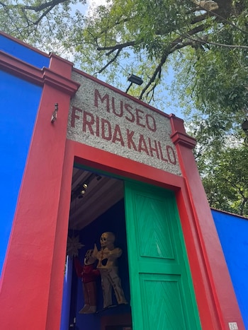 Fachada de un edificio azul y rojo con cartel 'MUSEO FRIDA KAHLO'. Puerta verde. Dos figuras esqueléticas dentro. Cielo y árboles en la parte superior
