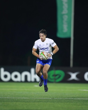 Un jugador de rugby con camiseta blanca y pantalón azul corre en un campo verde, sosteniendo un balón Gilbert. El fondo es oscuro con vallas publicitarias borrosas