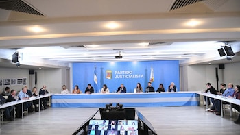 José Mayans y políticos en reunión. Al fondo, telón azul con logo del Partido Justicialista y bandera argentina. Pantalla en primer plano muestra videollamada