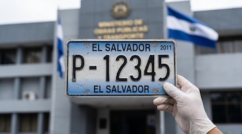 Mano enguantada sostiene una placa vehicular de El Salvador con letras negras y fecha 2011, sobre un edificio gubernamental gris con banderas nacionales de El Salvador.