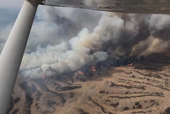 Vista aérea de un incendio forestal con denso humo blanco y gris elevándose sobre llamas anaranjadas en un terreno árido y montañoso. Parte del ala de un avión es visible
