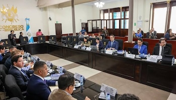 Personas vestidas de traje sentadas ante mesas con computadoras y micrófonos en una sala con el emblema del Congreso de Guatemala