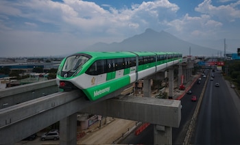 Tren monorriel verde y blanco de Metrorrey circula sobre una vía elevada de concreto. Debajo hay una carretera con autos y al fondo, montañas bajo un cielo nublado