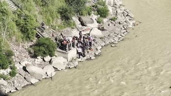 Un grupo de personas, algunas con chalecos salvavidas, se reúne en la orilla rocosa de un río junto a una estructura de escaleras metálicas