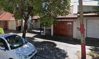 Vista exterior de una calle residencial con un auto blanco estacionado, un árbol frondoso y varias casas. Destaca una casa con portón de madera y reja oscura