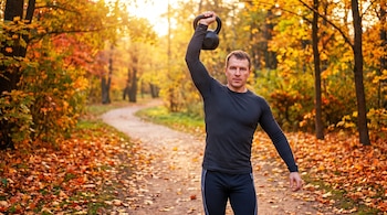 Hombre de mediana edad con ropa deportiva negra sostiene una pesa rusa sobre su cabeza. Fondo de parque otoñal con árboles, hojas anaranjadas y amarillas, y sendero.