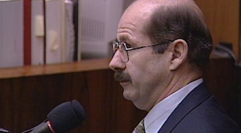 Un hombre de perfil con gafas, bigote y traje oscuro frente a un micrófono