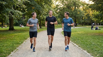 Tres hombres blancos, vestidos con ropa deportiva, corren por un sendero de tierra en un parque frondoso bajo la luz del sol.