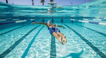 Vista submarina de una nadadora con gorro y traje azul, impulsándose en una piscina con carriles. La superficie del agua y palmeras son visibles.