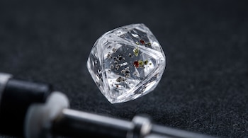 Un diamante transparente con inclusiones de partículas minerales negras, rojas y verdes flota sobre un fondo oscuro. Parte de una herramienta metálica desenfocada.