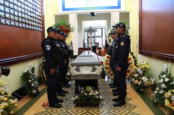 Dos agentes de policía en uniforme táctico cargan un ataúd gris adornado. Una fila de policías de uniforme blanco y azul saluda