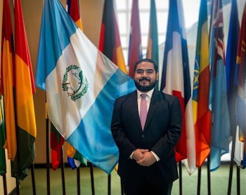 Hombre barbudo en traje oscuro y corbata rosa sonríe, de pie. Detrás, la bandera de Guatemala destaca entre otras banderas nacionales de diversos países