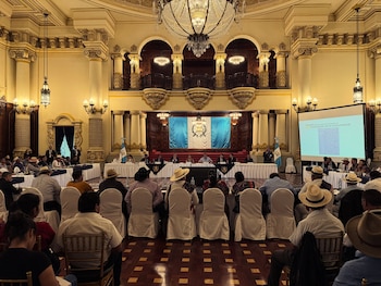 Salón ornamentado con columnas, candelabros y una audiencia de espaldas a la cámara. Al frente, una mesa con oradores y una pantalla proyectando información