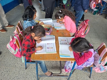 Vista superior de cuatro niñas en un pupitre de madera con cuadernos, lápices y mochilas. Se observan otras personas y un suelo de baldosas