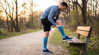 Un hombre viste camiseta azul y pantalón negro, ajusta medias de compresión azules con un pie en un banco de madera en un sendero de tierra.