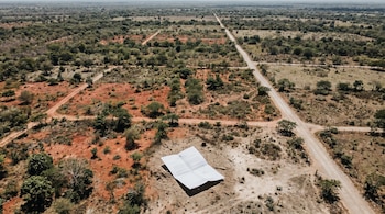 Vista aérea de un vasto terreno centroamericano con tierra rojiza y vegetación tropical. Caminos de tierra dividen el paisaje. Una hoja de papel oficial doblada yace en el suelo.