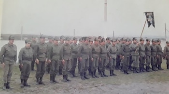 Una línea de soldados de infantería con uniformes militares verdes y gorras, de pie en formación al aire libre. Una bandera oscura está visible al fondo