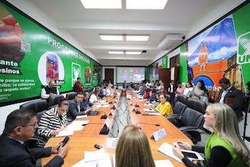 Personas sentadas alrededor de una larga mesa de madera con micrófonos y cuadernos en una sala decorada con paneles verdes