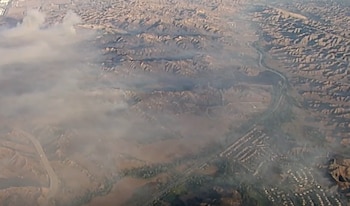 Vista aérea de un paisaje árido y montañoso cubierto por una gran extensión de humo gris, con carreteras y una zona residencial al fondo