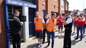 Un guardia de seguridad y policías detienen a dos hombres con chalecos naranjas en la entrada de Selhurst Park, mientras otros aficionados del Arsenal observan en un día soleado.