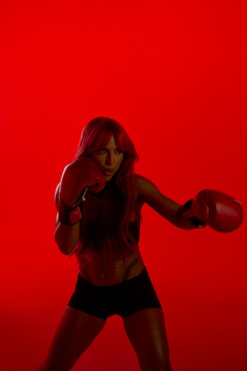Una mujer con cabello largo rojizo y guantes de boxeo rojos adopta una postura de combate sobre un fondo rojo vibrante, con una expresión concentrada