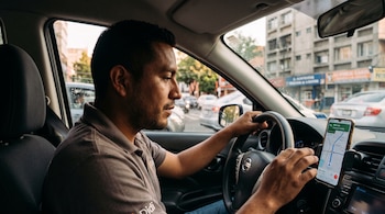 Un hombre frente al volante de un automóvil toca la pantalla de un teléfono inteligente sujeto al tablero que muestra un mapa de navegación.