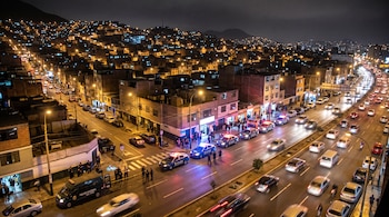 Una avenida urbana de noche con tráfico vehicular, patrullas de la Policía Nacional del Perú con luces encendidas y viviendas en cerros iluminados.