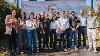 Doce personas sonrientes, hombres y mujeres, aplauden en un evento al aire libre. Detrás, una pantalla grande muestra el rostro de un hombre sonriente