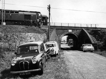 Vista en blanco y negro de una locomotora detenida en una vía elevada con personas a su lado, y coches estacionados en un camino de tierra debajo de un puente de ladrillo