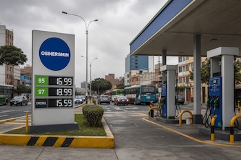 Un panel con el logotipo de Osinergmin y precios digitales junto a surtidores de combustible y vehículos en una calle con cielo nublado.