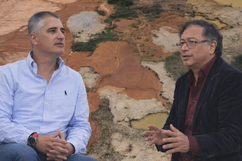 Dos hombres, uno en camisa azul claro y el otro en chaqueta oscura, conversan frente a un paisaje con terrenos áridos y formaciones de tierra.