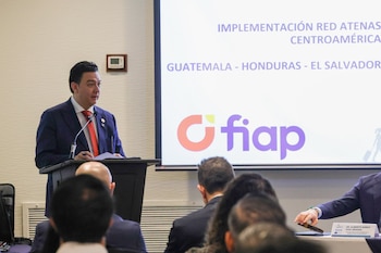 Personas sentadas en filas de sillas en un salón de conferencias frente a un podio, una pantalla blanca y banderas oficiales