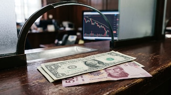Un billete de un dólar estadounidense y un billete de cincuenta pesos mexicanos sobre un mostrador de madera ante la pantalla de un monitor.