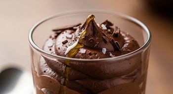 Primer plano de mousse de chocolate oscuro en un cuenco de vidrio, con un chorro de aceite de oliva, escamas de sal y virutas de chocolate encima.